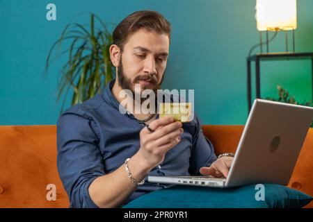 Jeune homme libanais utilisant une carte de crédit et un ordinateur portable tout en transférant de l'argent, achats en ligne, commande de livraison de nourriture. Portrait d'un homme arabe à la maison appartement salon sur canapé Banque D'Images