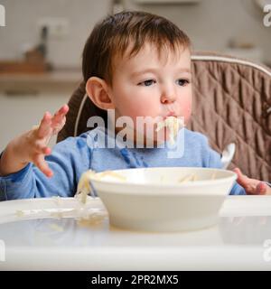 Un enfant drôle mange une pomme râpée avec sa bouche pleine tout en étant assis sur une chaise de cuisine. Bébé garçon affamé, il a de la nourriture dans sa bouche, de l'humour. Enfant âgé Banque D'Images