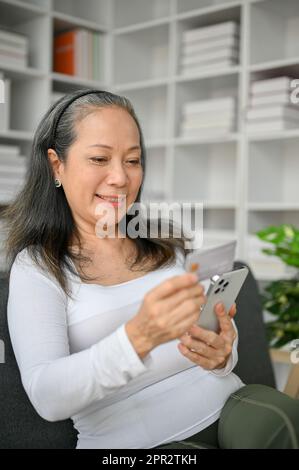 Bonne femme âgée faisant des achats en ligne avec un smartphone en utilisant une carte de crédit tout en étant assise sur le canapé. Banque D'Images