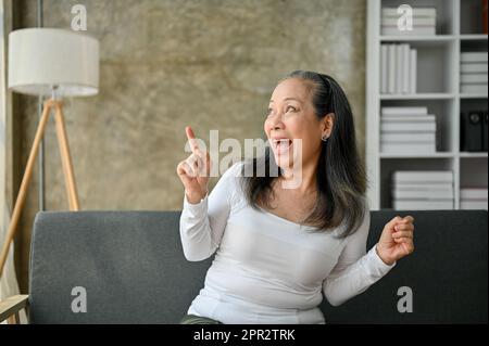Une femme asiatique mûre surjoyeuse et stupéfaite s'assoit sur un canapé dans son salon, regarde au loin et pointe son doigt de côté. Banque D'Images