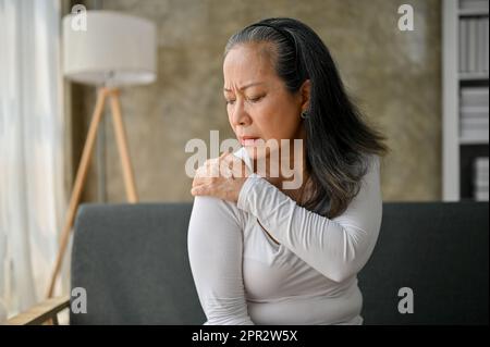 Malheureuse femme asiatique mature souffrant de douleurs à l'épaule, massant son épaule tout en se reposant sur un canapé dans son salon. Banque D'Images