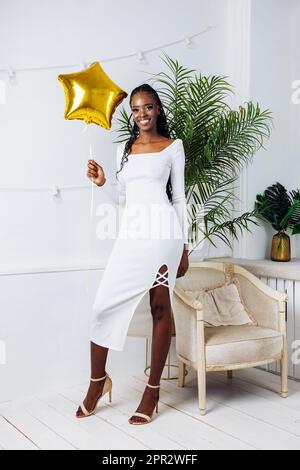 Souriante, élégante, brillante femme américaine moderne africaine en robe blanche bodycon tenir l'étoile d'or ballon sur la fête de vacances Banque D'Images