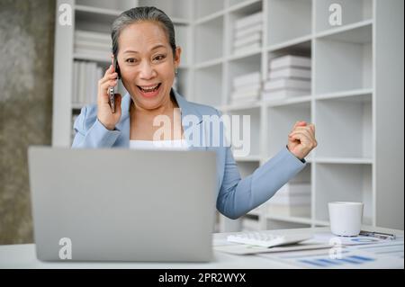 Heureuse et joyeuse femme d'affaires senior se sentant sortie après avoir entendu une bonne nouvelle de l'appel téléphonique au travail. Banque D'Images