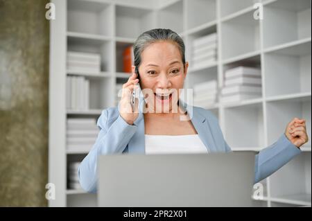 Heureuse et surjoyeuse femme d'affaires senior se sentant sortie après avoir entendu une nouvelle de dieu de l'appel téléphonique au travail. Banque D'Images