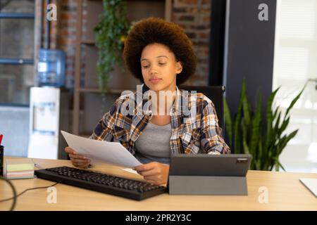 Bonne femme d'affaires décontractée biraciale assise au bureau et lisant le document Banque D'Images