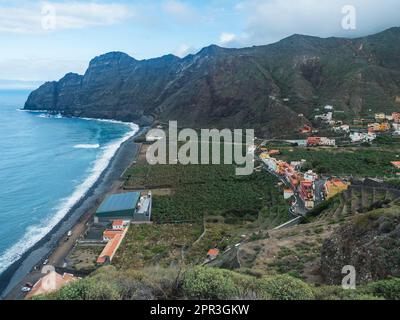 Vue aérienne de la plage de galets rocheux noirs de Playa Santa Catalina et du village d'Hermigua avec des maisons colorées et des plantations de bananes, des falaises de montagne Banque D'Images