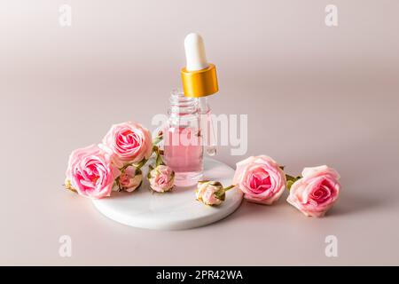 Une bouteille ouverte remplie d'une pipette d'un produit cosmétique naturel à base d'huile de pétale de rose se dresse sur un podium en marbre blanc avec des fleurs de rose Banque D'Images