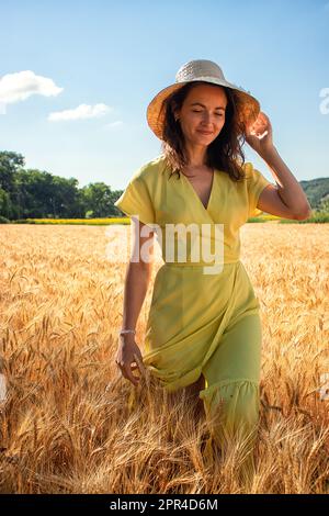 une jeune femme de race blanche, vêtue d'une robe jaune avec un chapeau de paille sur sa tête, se promette volontiers à travers un champ de blé, à la périphérie du village Banque D'Images