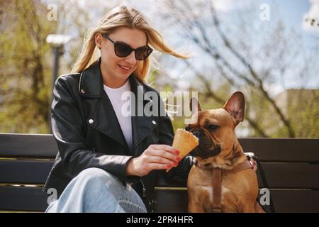 Belle jeune femme blonde assise sur un banc et nourrissant un chien avec de la crème glacée. Adorable petit chiot bulldog qui apprécie le dessert Banque D'Images