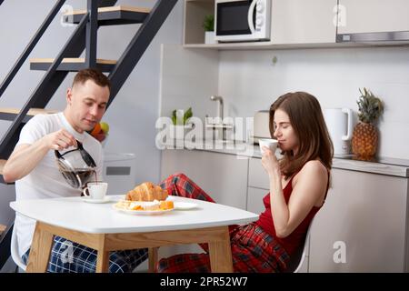 Petit déjeuner coziness. Jeune couple heureux, homme et femme en pyjama ayant le petit déjeuner dans la cuisine, boire du café, parler Banque D'Images