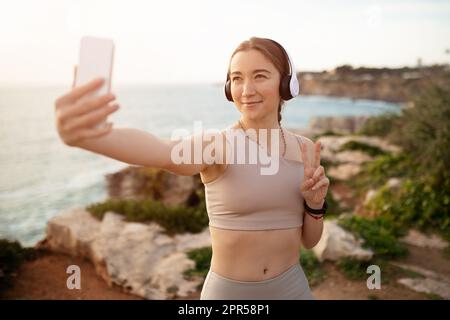 Jeune femme caucasienne gaie dans les vêtements de sport et les écouteurs sans fil faire selfie sur le téléphone Banque D'Images