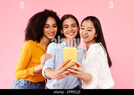 Joyeuses femmes diverses faisant le selfie sur téléphone portable, debout ensemble sur fond rose studio Banque D'Images
