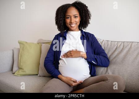 Souriante jeune afro-américaine cally femme enceinte dans une ambiance décontractée s'assoit sur le canapé touche le gros ventre avec les mains Banque D'Images