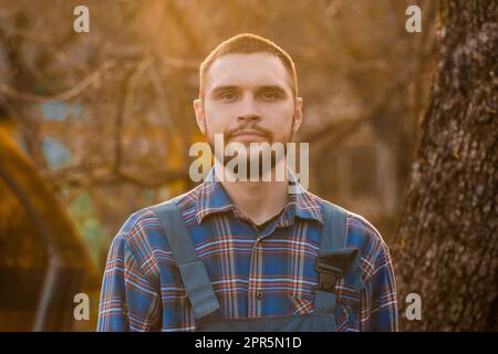 Un fermier d'apparence européenne portrait rural en plein soleil coucher de soleil avec une barbe, une chemise et une combinaison regarde l'appareil photo à l'extérieur. Banque D'Images