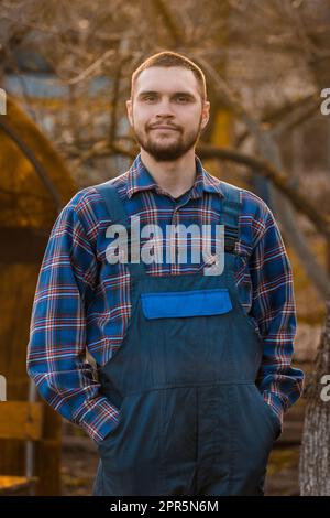 Fermier sourire européen portrait rural mâle avec barbe, chemise et salopette avec les mains dans les poches regardant l'appareil photo à l'extérieur coucher du soleil. Banque D'Images