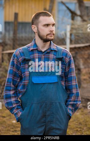 Fermier beau portrait rural d'apparence européenne avec barbe, chemise et salopette avec les mains dans les poches donnant sur la campagne caoutdoor. Banque D'Images