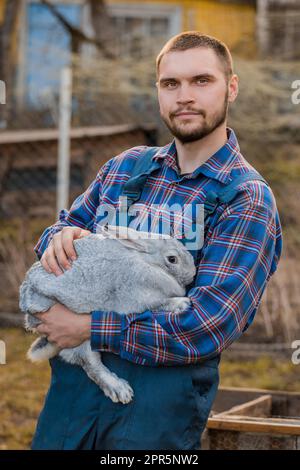 Un fermier fait un beau portrait rural européen du caucase en campagne avec une barbe, une chemise et une combinaison regardant la caméra avec un lapin blanc dans ses bras outdoo Banque D'Images