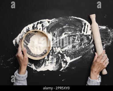 Les mains des femmes tiennent un tamis rond en bois avec de la farine de blé blanc sur une table noire, vue de dessus Banque D'Images