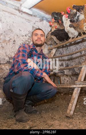 Un fermier d'apparence européenne, avec une barbe, en combinaison, regarde la caméra à côté des poules dans la grange. Volaille. Banque D'Images