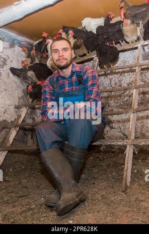 Un fermier content d'apparence caucasienne, jeune, beau, avec une barbe dans une chemise et des salopettes à côté d'une perche pour les poulets. Volaille et ménage. Banque D'Images