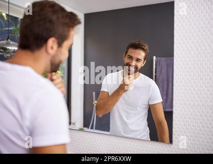 Mais j'ai l'air bien avec une barbe. Photo courte d'un beau homme se regardant dans le miroir tout en touchant sa barbe à la maison. Banque D'Images