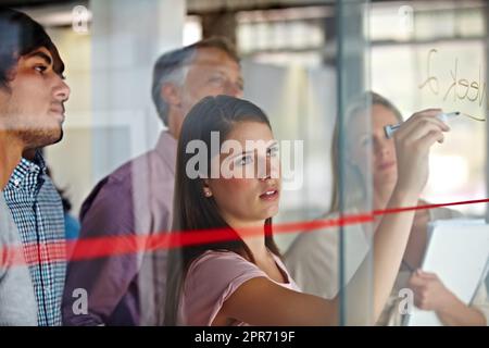 Expliquer ses idées avec un dessin. Une jeune femme d'affaires écrit ses plans sur un verre tandis que ses associés regardent. Banque D'Images