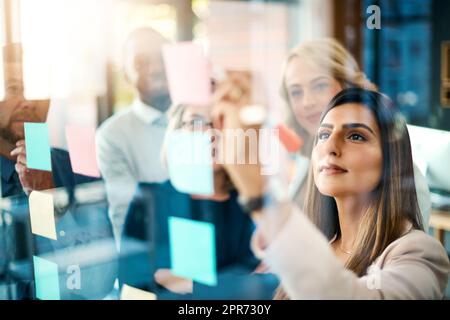 La planification est toujours une étape cruciale dans la réussite. Photo d'un groupe de femmes d'affaires qui organisent des notes adhésives sur un mur de verre dans un bureau moderne. Banque D'Images