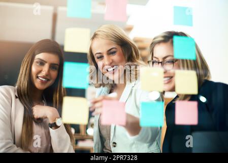 Brainstorming leur manière de faire des affaires brillantes. Photo d'un groupe de femmes d'affaires qui organisent des notes adhésives sur un mur de verre dans un bureau moderne. Banque D'Images