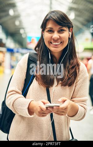 J'ai reçu mon ticket et je suis prêt à me lancer. Portrait rogné d'une jeune femme attrayante écoutant de la musique tout en marchant dans une station de bus. Banque D'Images