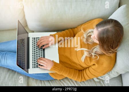 Connecté au monde depuis le confort de la maison. Prise de vue en grand angle d'une jeune femme utilisant son ordinateur portable tout en étant assise sur le canapé à la maison. Banque D'Images