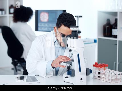 Un scientifique médical sérieux assis à un bureau et utilisant un microscope pour examiner et analyser des échantillons de test sur des lames. Professionnel de la santé hispanique découvrant un remède aux maladies dans son laboratoire Banque D'Images