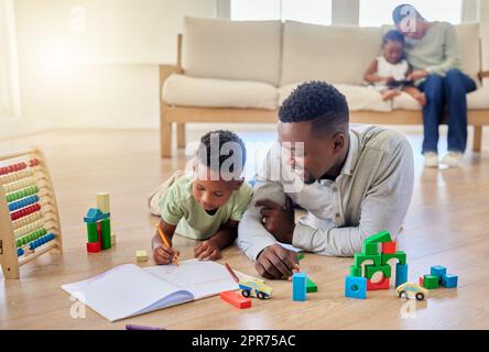 Jeune père afro-américain heureux aidant son fils à faire ses devoirs tout en étant assis sur le sol à la maison. Un petit garçon s'est concentré sur l'apprentissage et la réalisation d'une tâche. Dessin d'un petit garçon dans un livre Banque D'Images