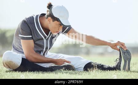 Longueur totale d'un joueur de baseball assis sur un terrain et s'étirant avant de jouer à un match. Un athlète sérieux et concentré se prépare à jouer sur l'herbe. Homme en forme, actif, sportif, sportif Banque D'Images