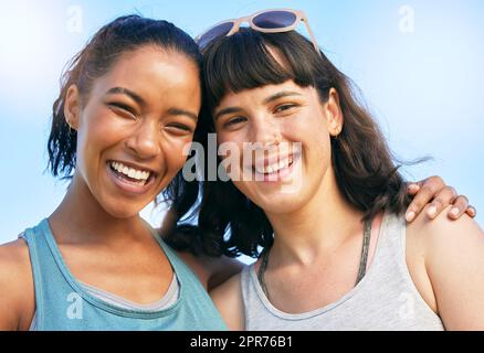 Portrait de deux amis souriants s'embrassant tout en se tenant debout contre le ciel bleu. Souriante et heureuse, les jeunes femmes se colent et s'embrasent à l'extérieur pendant les vacances d'été. Fermez les meilleurs amis qui s'amusent le week-end Banque D'Images