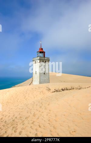 Phare déserté au bord de la mer avec ciel bleu en arrière-plan par une journée ensoleillée. Un phare entre la zone sablonneuse entourée d'eau et ciel nuageux. La vieille tour mystérieuse seule dans le désert Banque D'Images