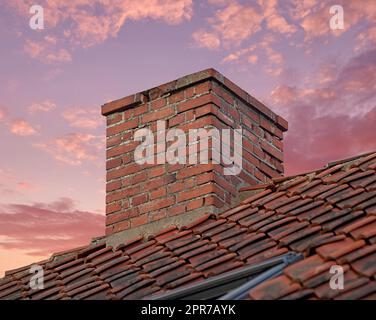 Gros plan d'une cheminée en briques rouges contre un ciel de coucher de soleil coloré pour les gaz de combustion et l'isolation de la maison sur le toit carrelé. Conception architecturale sur le bâtiment de la maison pour l'extraction de fumée de la cheminée ou du four Banque D'Images