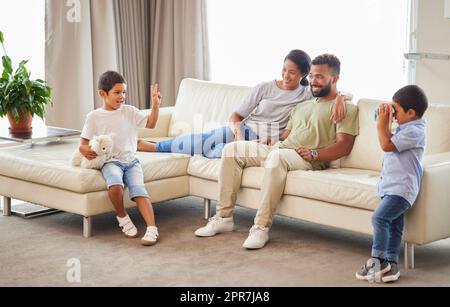 Jeunes familles mixtes se liant dans le salon pendant que les parents se détendent sur le canapé pendant que leurs deux petits fils jouent. Couple hispanique souriant tout en regardant leurs garçons jouer dans le salon Banque D'Images