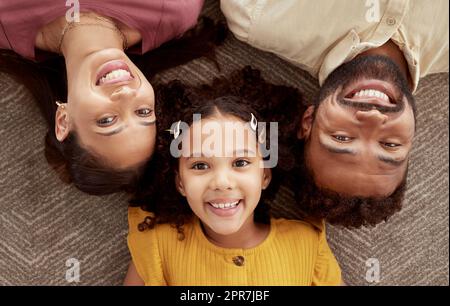 Au-dessus du portrait des parents de race mixte appréciant le week-end avec la fille dans le salon à la maison. Fille hispanique souriante embrassant la mère et le père et se liant dans le salon. Couple heureux allongé avec l'enfant Banque D'Images