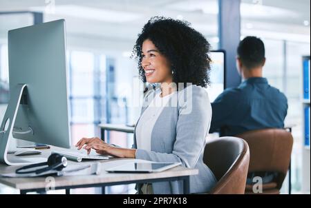 Un jeune heureux agent de télémarketing de centre d'appels de course mixte parlant sur un micro-casque tout en travaillant sur un ordinateur dans un bureau. Service d'assistance téléphonique féminin fiable et convivial pour le service clientèle et l'assistance commerciale Banque D'Images