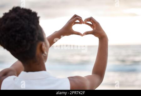 Vision arrière des mains sur une femme afro-américaine en train de faire un coeur contre le ciel à l'extérieur. Femme noire confiante debout sur une plage à l'extérieur au coucher du soleil se faisant un geste vers la mer. Santé et amour Banque D'Images