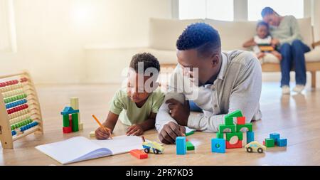 Jeune père afro-américain heureux aidant son fils à faire ses devoirs tout en étant assis sur le sol à la maison. Petit garçon s'est concentré sur l'écriture dans un bloc-notes et de faire une tâche. Dessin d'un petit garçon dans un livre Banque D'Images