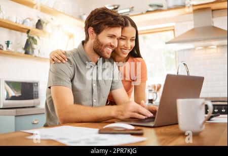 Jeune couple interracial gai se liant tout en travaillant sur un ordinateur portable ensemble à la maison. Petit ami et petite amie caucasiens travaillant à travers leurs factures et utilisant un ordinateur portable. Contenu épouse et mari naviguant en ligne sur un ordinateur Banque D'Images