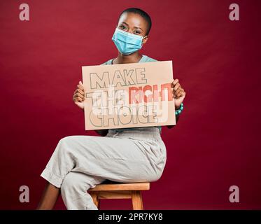 Femme vaccinée Covid afro-américaine montrant, tenant une affiche et portant un masque chirurgical. Modèle noir heureux isolé sur fond rouge studio avec espace de copie. Promotion du vaccin corona avec signe Banque D'Images