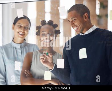 Trois hommes d'affaires heureux écrivant des idées et brainstorming ensemble dans un bureau au travail. Homme d'affaires hispanique gai faisant une note sur une fenêtre en verre avec des collègues féminins Banque D'Images