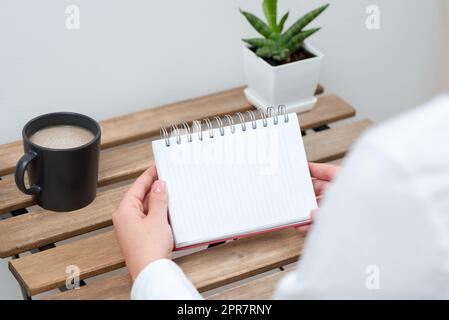 Femme tenant un cahier avec message important sur la table avec café et plante. Femme d'affaires présentant des informations de base sur le bureau avec une coupe et une fleur. Banque D'Images