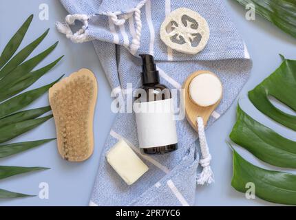 Une bouteille en verre à pompe sur une serviette de bain bleue près des feuilles tropicales et des accessoires de soin de la peau, maquette Banque D'Images