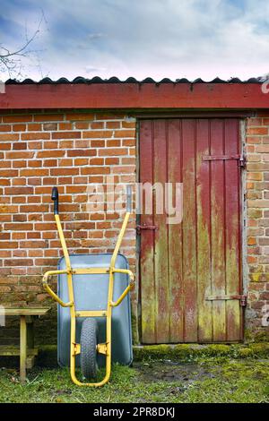 Une photo d'une brouette à l'envers, d'une petite ferme en briques avec une porte rouge et jaune mélangée, et d'une table en bois sur de l'herbe humide. Une belle photo d'une petite ferme sous le ciel lumineux. Banque D'Images