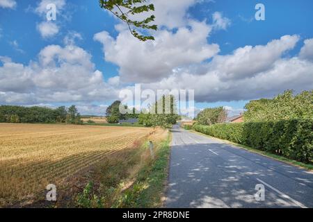 Paysage rural dynamique d'une route à travers la campagne par une journée ensoleillée. Champ sec de maïs ou de blé après récolte dans un petit village danois, dans un ciel bleu nuageux. Calme ville agricole en été Banque D'Images