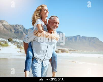 Couple d'âge mûr heureux en vacances sur la plage. Mari senior actif offrant à sa femme une promenade en pigeyback tout en profitant d'une journée ensoleillée à l'extérieur. Homme et femme énergiques s'amusant pendant les vacances Banque D'Images