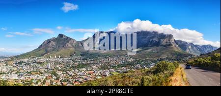 Une route de montagne donnant sur la ville avec un ciel bleu nuageux. Paysage panoramique de montagnes verdoyantes entourant une ville urbaine et une route pittoresque pour voyager le long du Cap, Afrique du Sud Banque D'Images